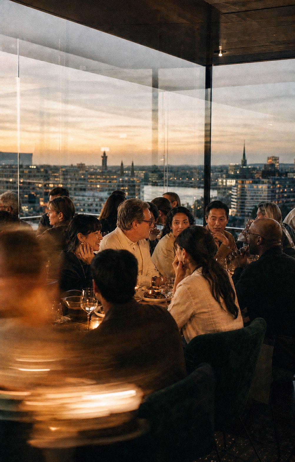 Members at a private candlelit dinner high above a city skyline at dusk, in conversation.